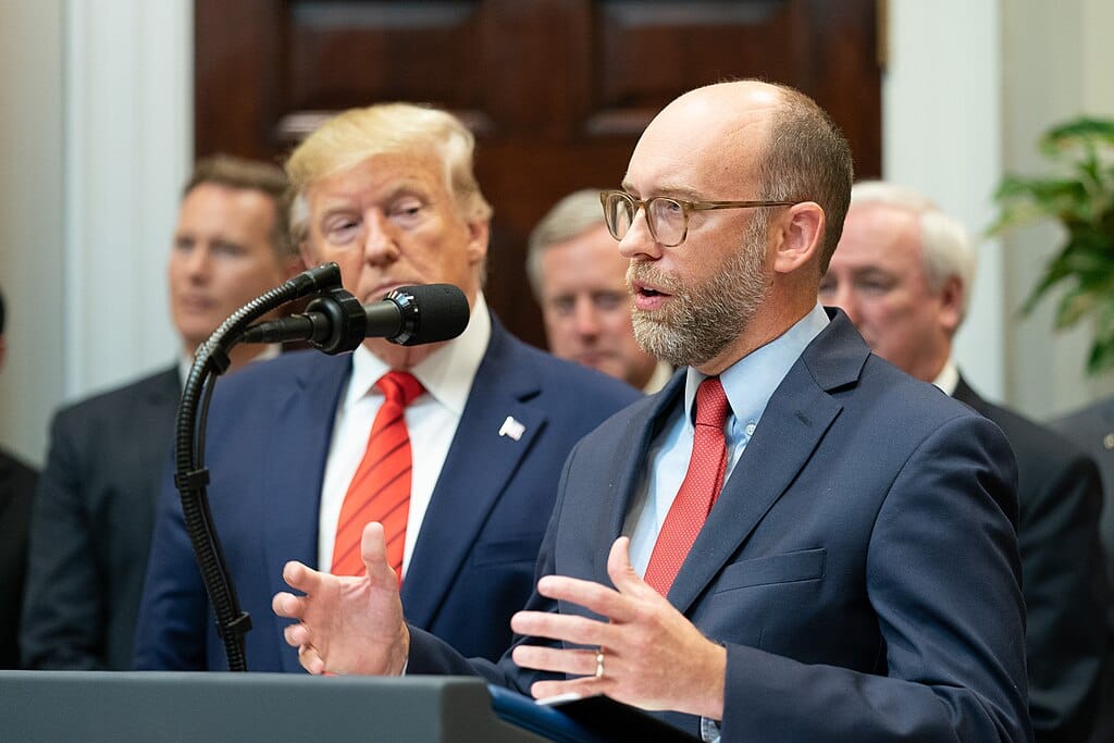 Russell Vought speaks at a podium with Trump next to him and other men behind them