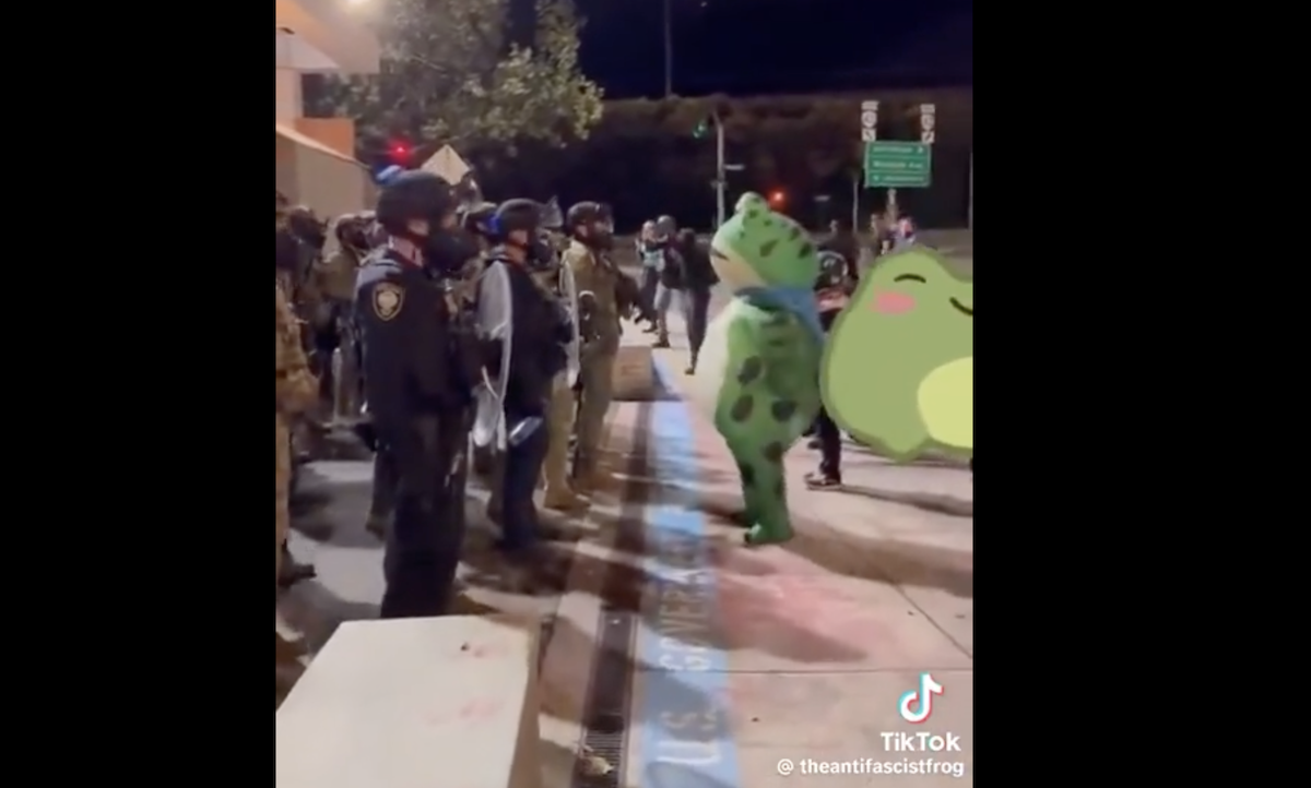 Some troops stand with shields and masks in Portland while a person in an inflatable frog costume thrusts at them.