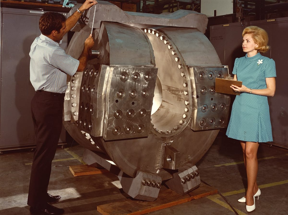 A man and a woman stand next to a large metal structure used in a fusion experiment, making measurements, in the 1960s