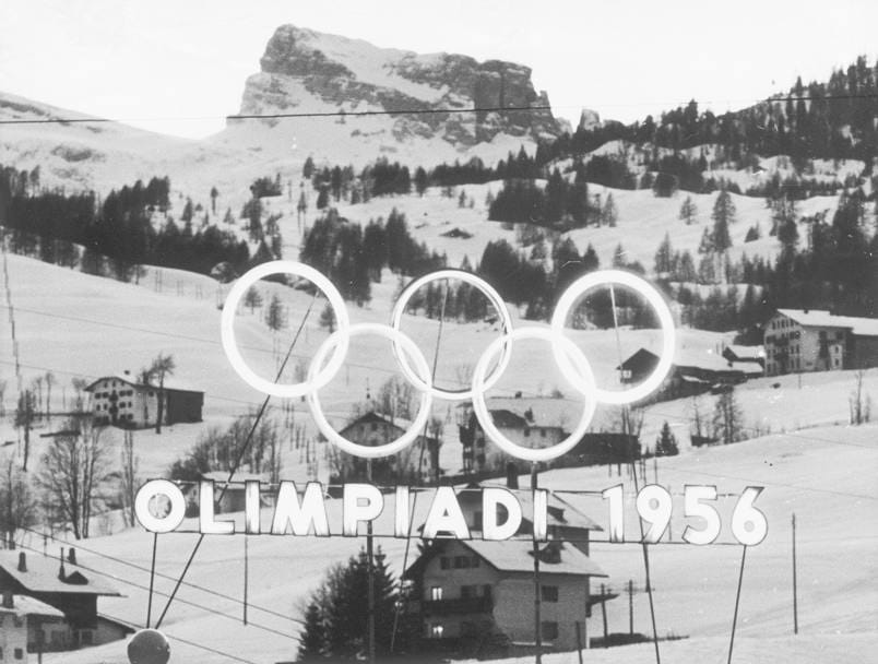 The Olympic rings over text reading "Olimpiadi 1956" in front of a snowy mountain in Cortina, Italy