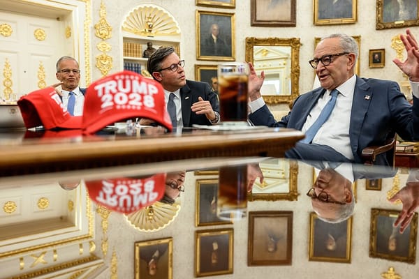 Chuck Schumer sitting at a table in the White House, gesticulating, with John Thune and Mike Johnson next to him and a "Trump 2028" hat on the table