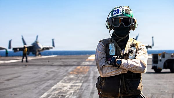 A Navy sailor with a full black mask and goggles on, on the deck of an aircraft carrier, with fighter jets in the background